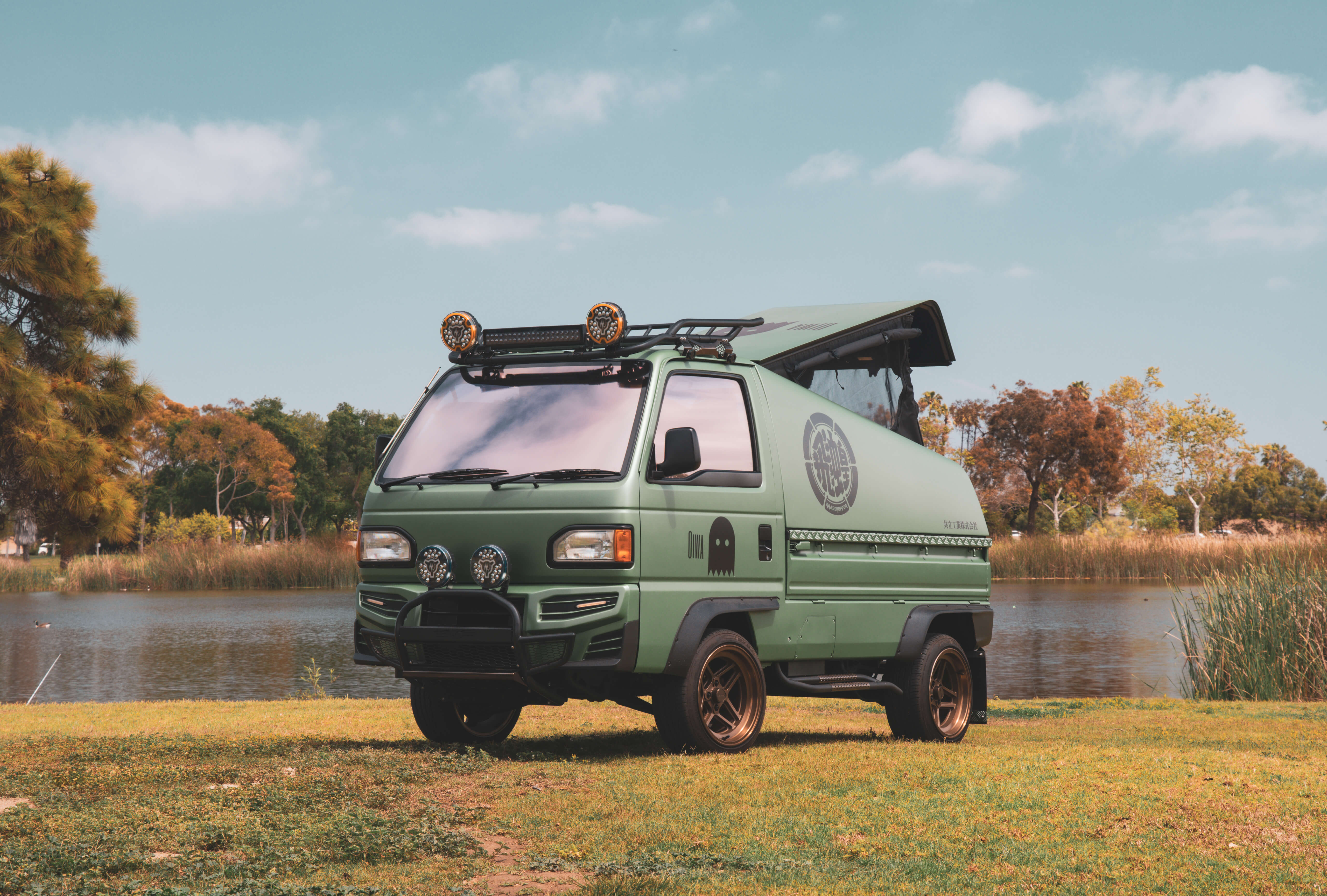 Green Honda Acty Grasshopper kei truck camper with pop-up metal camper shell, lifted suspension, roof rack, and off-road lighting parked in scenic outdoor setting.