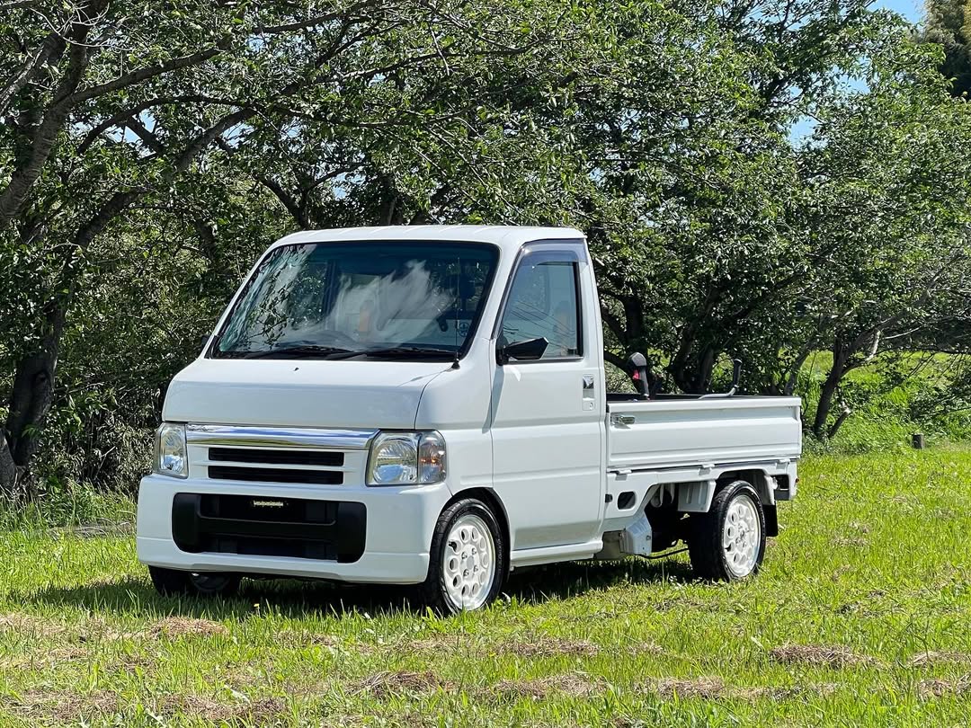 White Honda Acty HA6, HA7 (1999-2009) kei truck parked on grassy terrain, featuring aftermarket white wheels, clean styling, and highlighting its versatility.