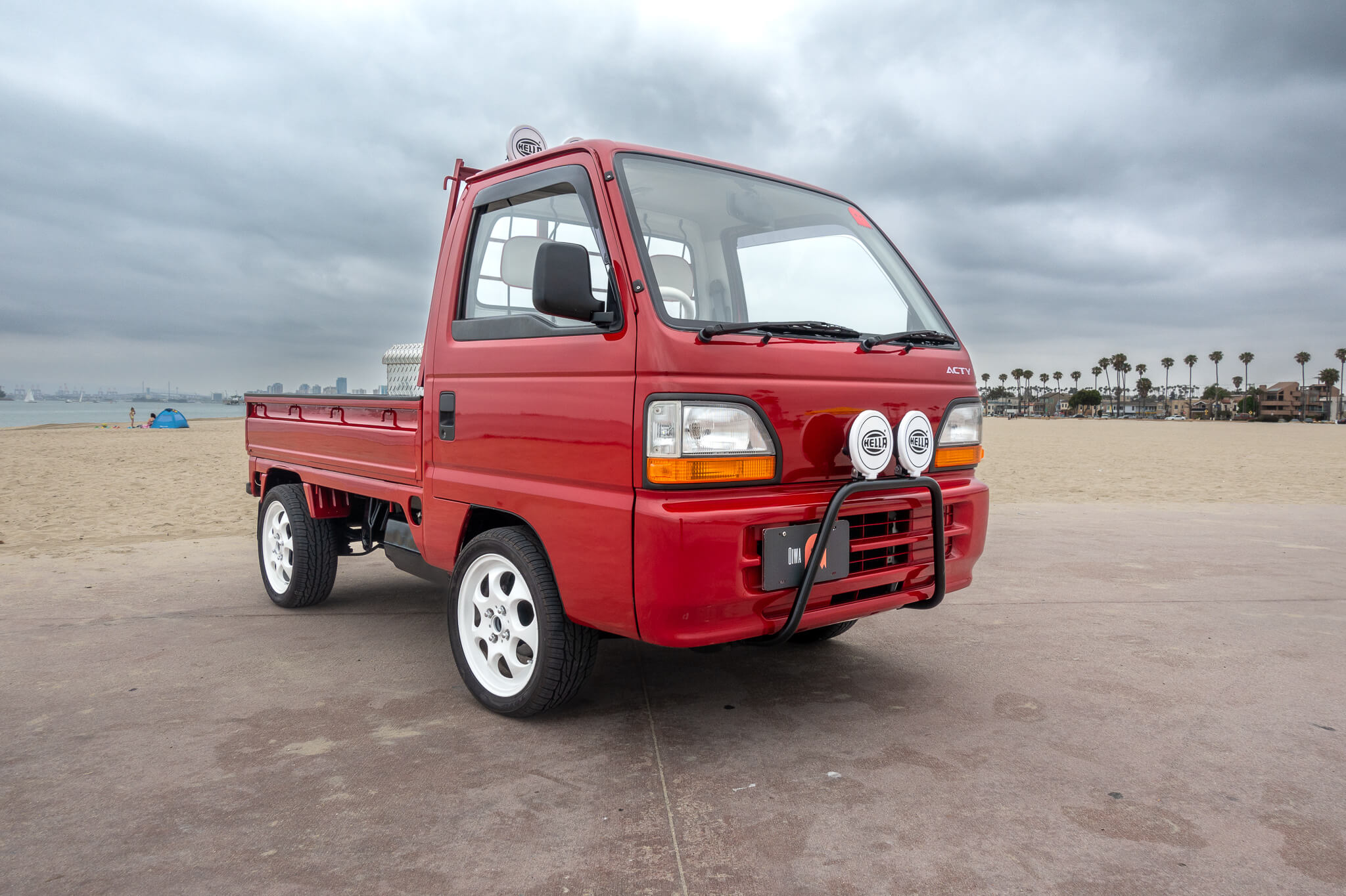 Red Honda Acty Truck HA3, HA4 (1990-1999) with white aftermarket wheels, front brush guard, and HELLA fog lights, parked on a beach showcasing its custom style.