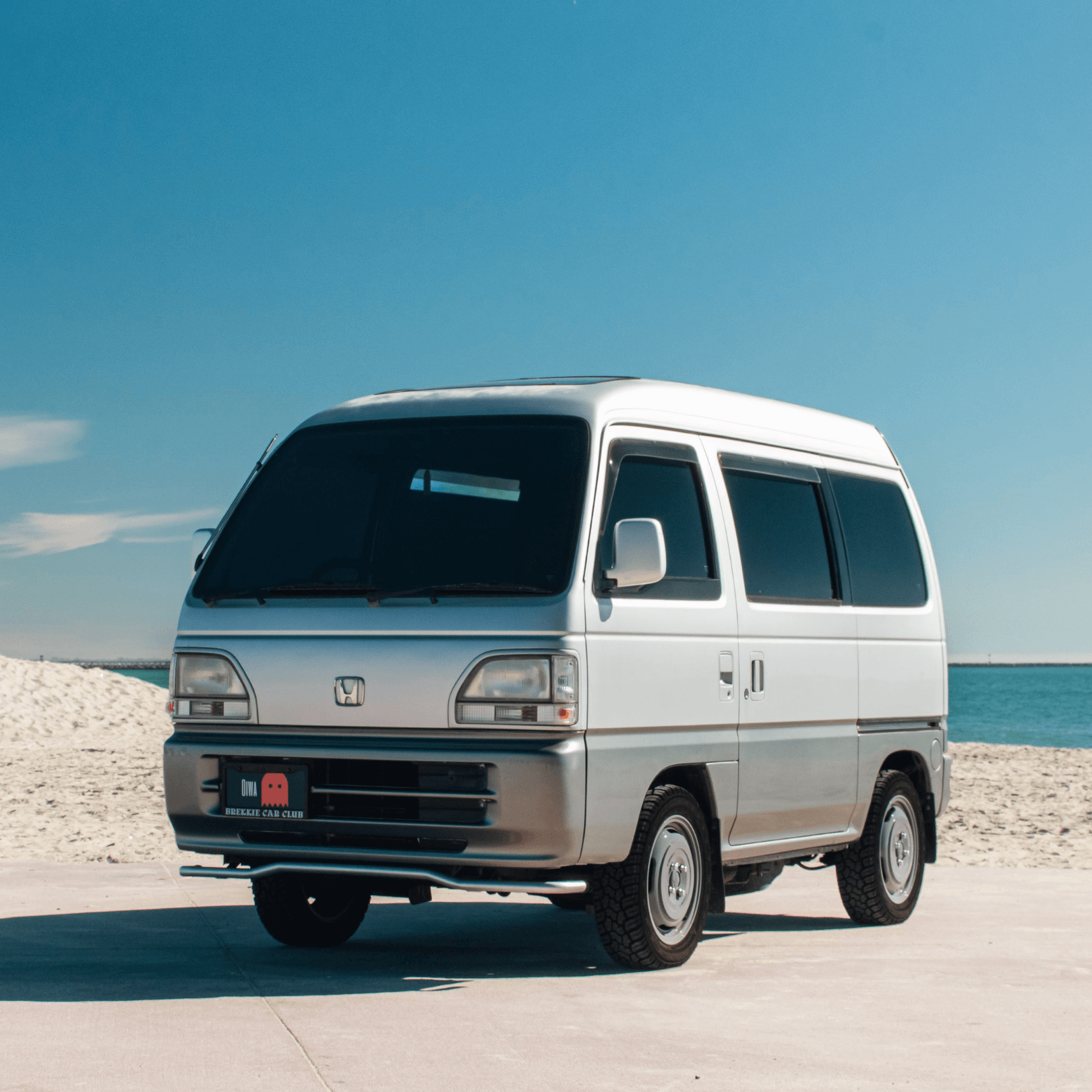 Grey and silver Honda Acty Van HH3, HH4 (1990-1999) parked at a beach, emphasizing its compact size, retro styling, and practicality for leisure and urban use.